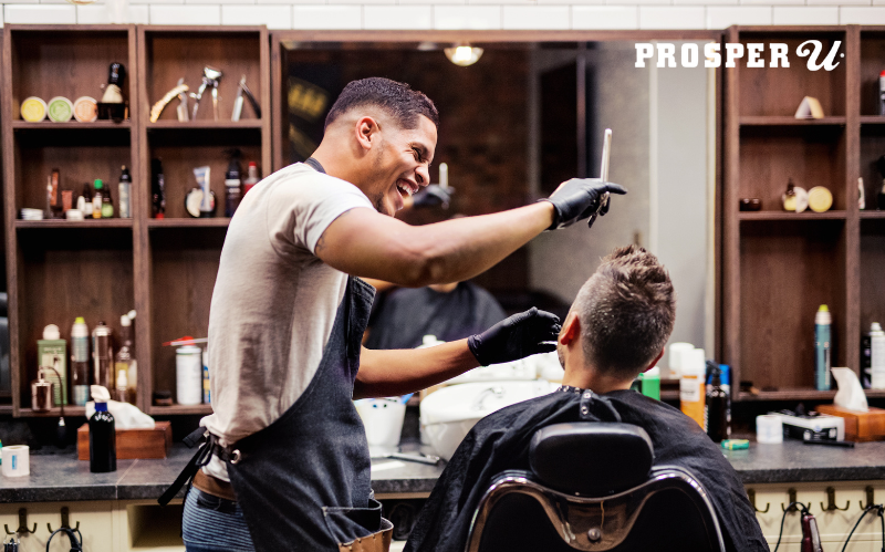 Black barber laughing with a client who is sitting in their chair. They are having a great conversation.